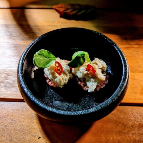 Two small tapas in a small black bowl on a wooden table in an elegant restaurant.